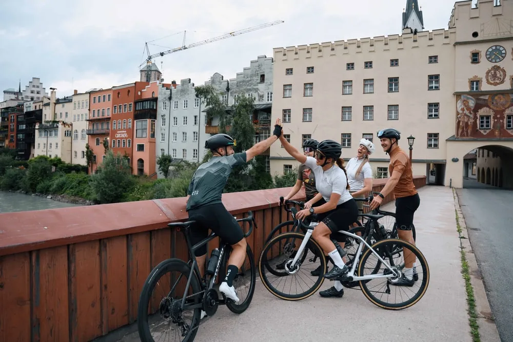 A group of cyclists high-fiving on a bridge with Wasserburg am Inn's colorful historic buildings in the background.