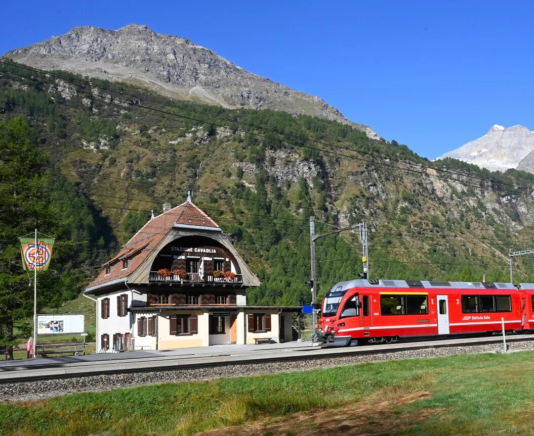 A red Rhaetian Railway train passes the charming Stazione Cavaglia building beneath dramatic Alpine peaks.
