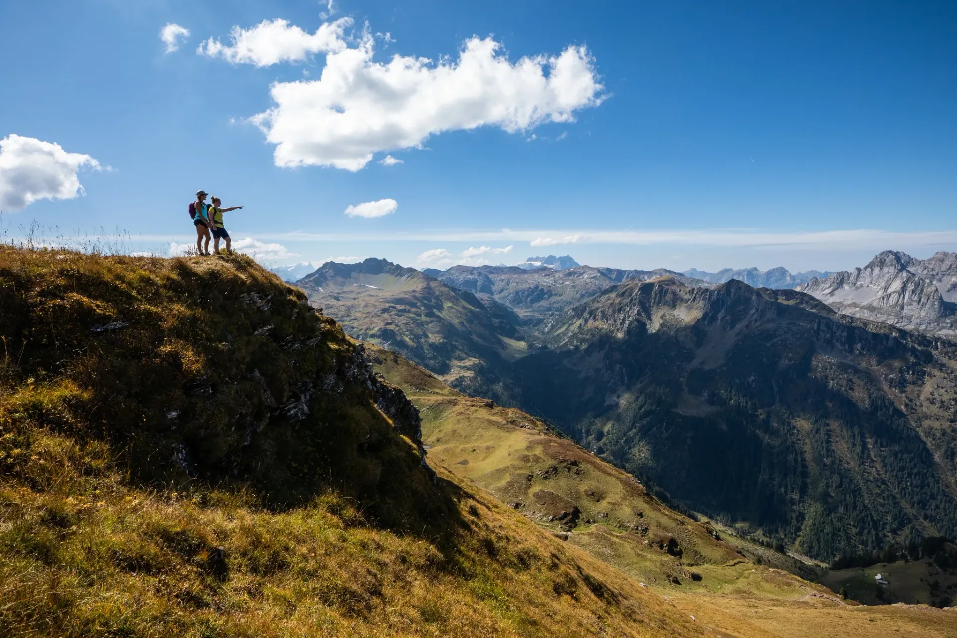 Two hikers stand on a rocky alpine summit overlooking a sweeping mountain valley under blue skies.