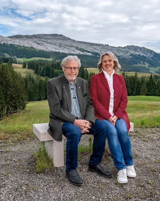 Two hotel owners seated on a stone bench with panoramic Alpine meadow backdrop behind them.
