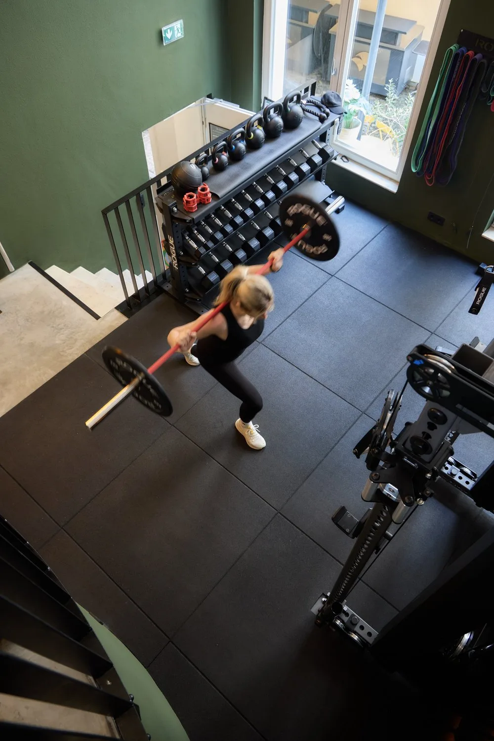 A woman performs a barbell deadlift in a well-equipped hotel gym with Rogue dumbbells and kettlebells.