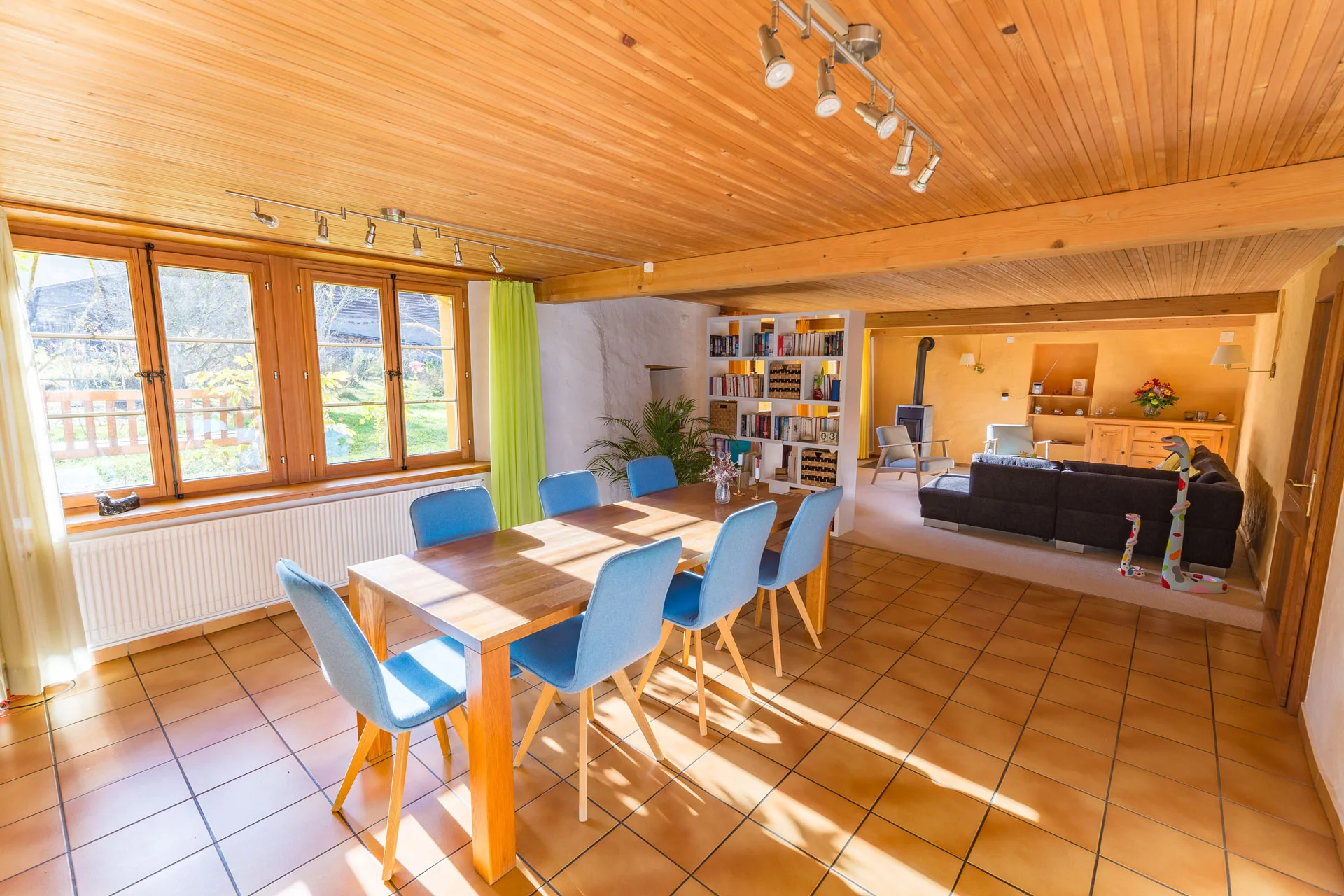 Sunlit open-plan dining and living area with wooden ceiling, blue chairs, and bookshelf beyond.
