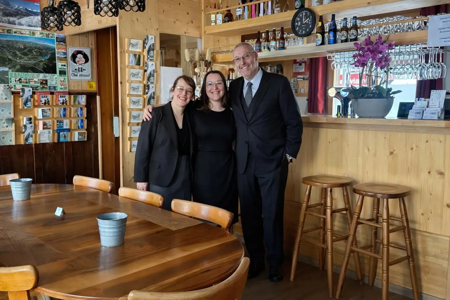 Three smiling staff members in formal black attire pose warmly inside the cozy wooden-interior restaurant.