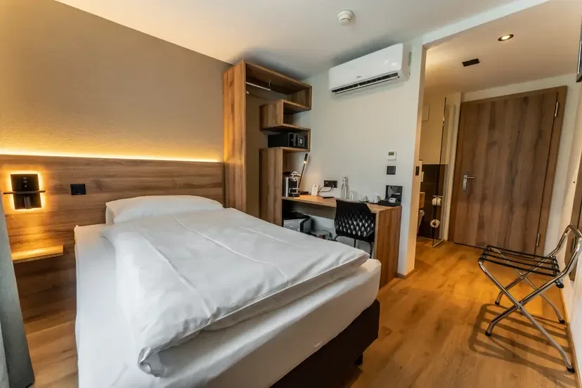 A clean, functional single hotel room with wood furnishings, a work desk, and warm bedside lighting.