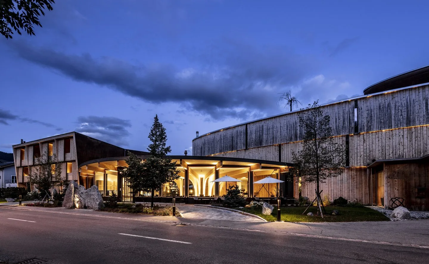 Warmly lit wooden curved building with outdoor terrace and mountain backdrop at dusk.