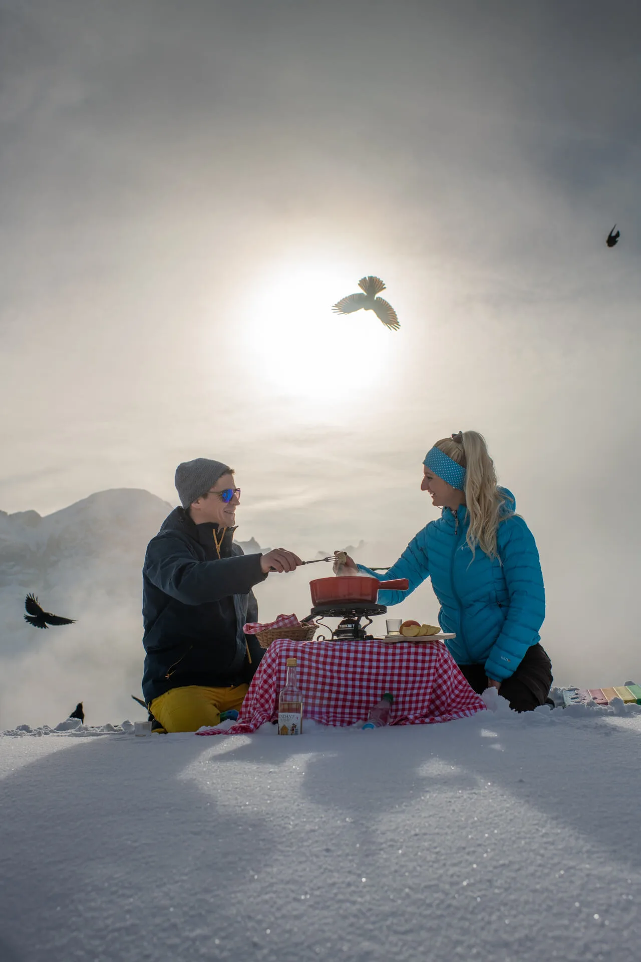 A couple enjoys a romantic fondue picnic in the snow at altitude with alpine choughs flying around them.